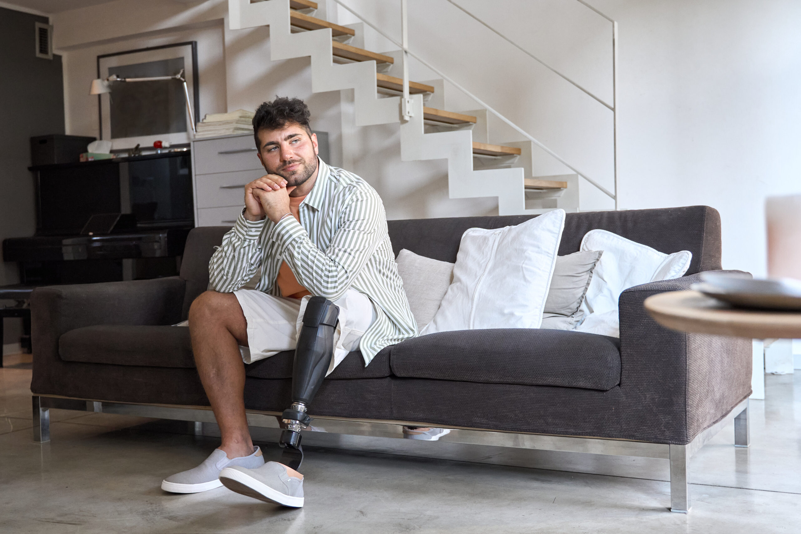 Young thoughtful amputee man with leg prosthesis sitting on sofa.